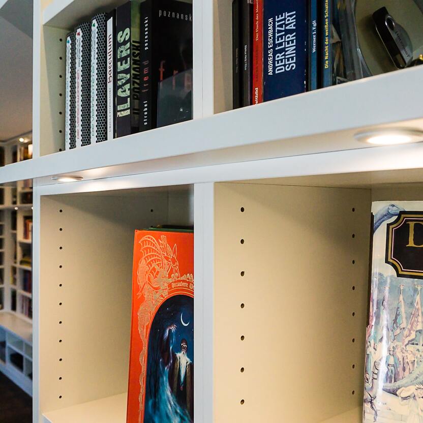 Maßgefertigte Bücherwand mit flexibel anpassbaren Fächern Detailaufnahme einer individuell angefertigten Bücherwand mit höhenverstellbaren Regalböden und integrierter LED-Beleuchtung, gefüllt mit Büchern und Deko-Elementen.