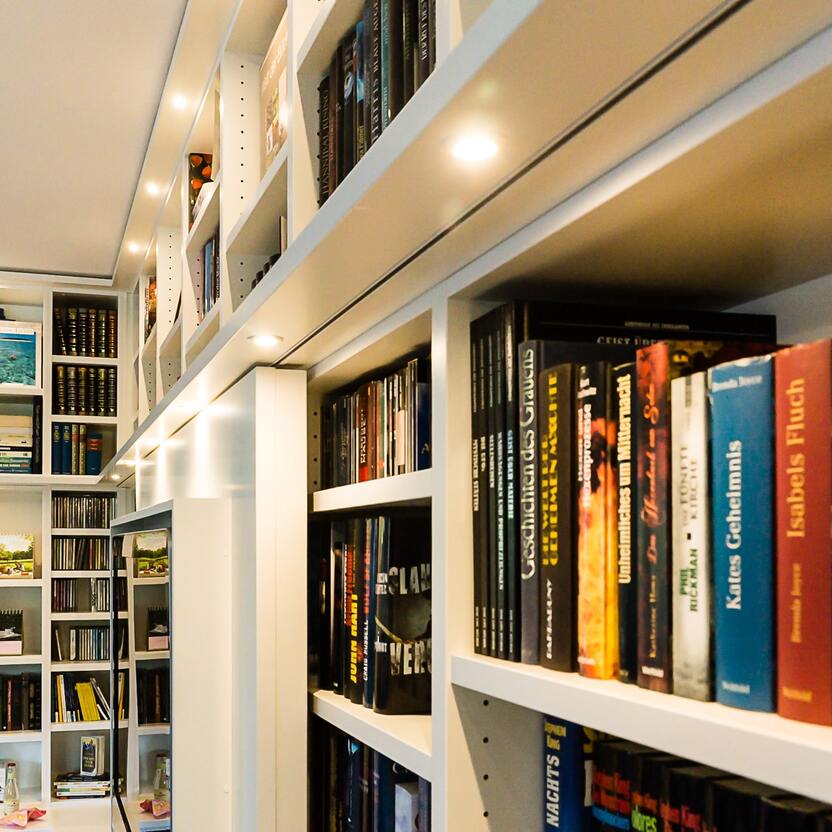 Maßgefertigte Bücherwand mit LED-Beleuchtung Detailansicht einer individuell angefertigten Bücherwand mit integrierter LED-Beleuchtung, gefüllt mit Büchern und Medien für eine stilvolle Wohnraumgestaltung.