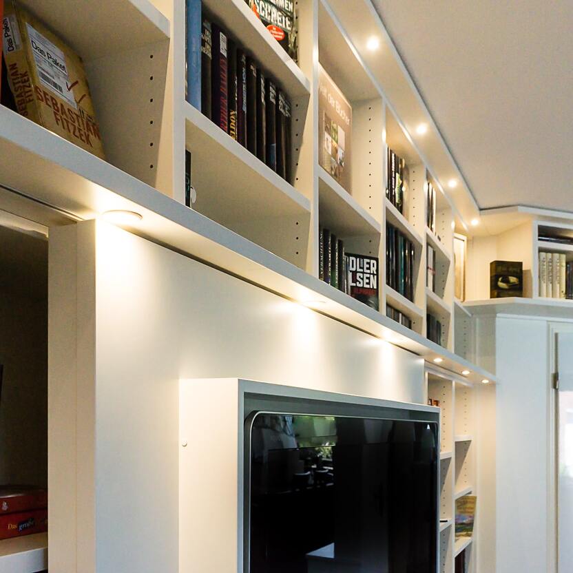 Modernes Bücherregal mit integrierter Beleuchtung Großes, weißes Bücherregal mit eingebauter Beleuchtung, gefüllt mit verschiedenen Büchern und einem Fernseher, in einem stilvoll eingerichteten Raum.