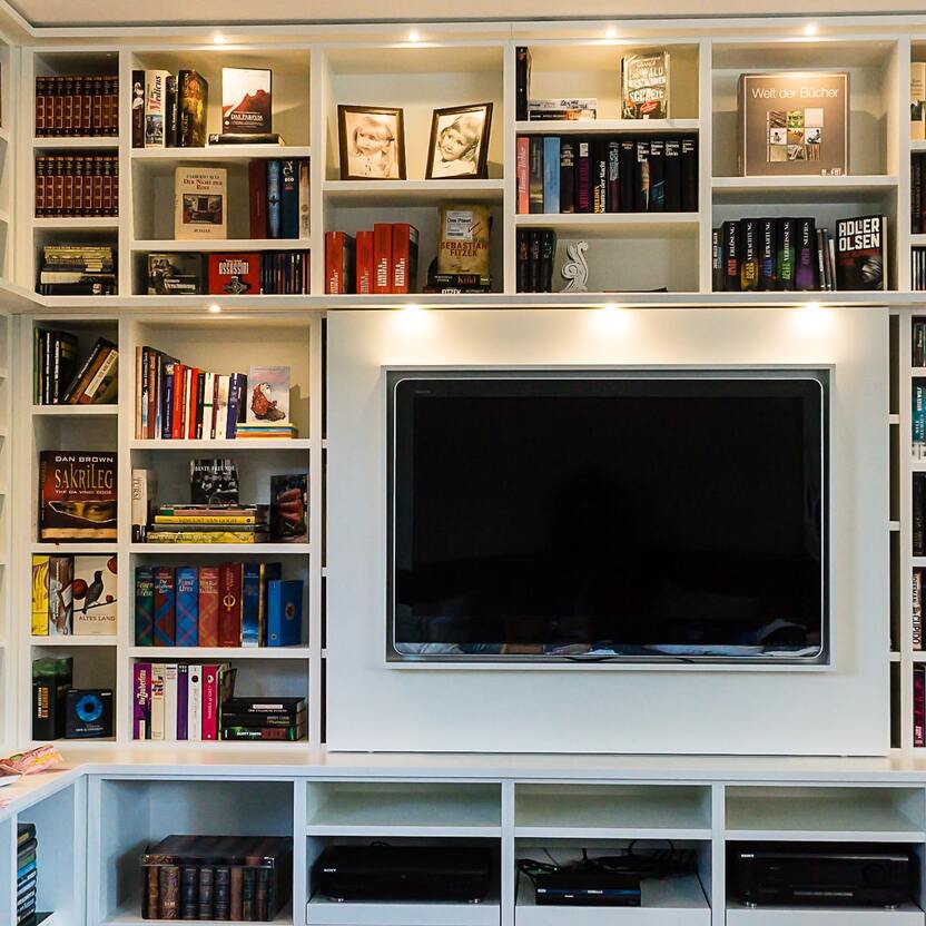 Modernes Mediencenter mit umfangreicher Büchersammlung Breites, weißes Regalsystem mit integriertem Fernseher und vielfältigen Büchern, darunter Klassiker und moderne Literatur, in einem gemütlichen Raum.