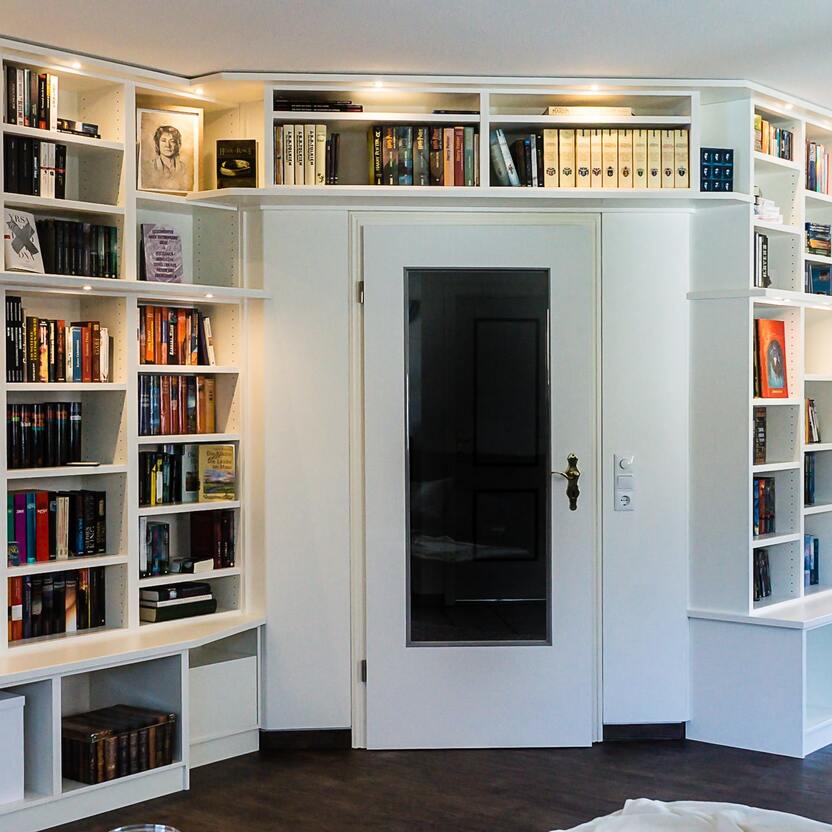 Wohnzimmer mit eingebauten Bücherregalen um eine Tür herum Ansicht eines Wohnzimmers mit weißen, bodenbis deckenhohen Bücherregalen, die eine zentrale Tür umrahmen, reich bestückt mit einer Vielzahl von Büchern.