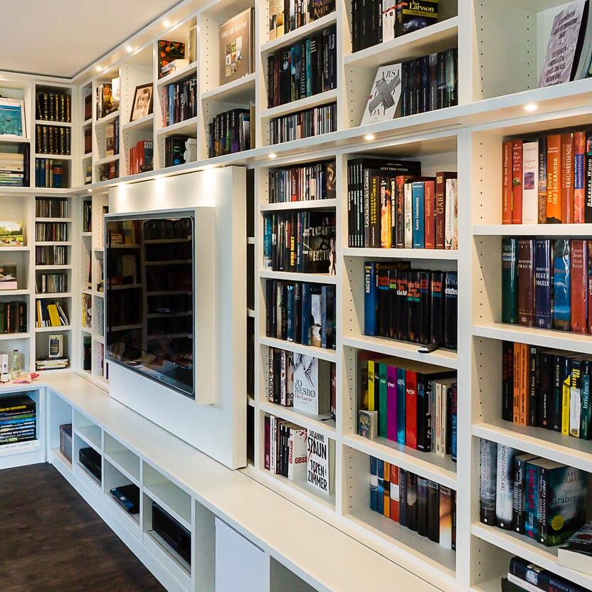 Umfangreiches Bücher- und Mediaregal in einem Wohnzimmer Helles, modernes Regalsystem in einem Wohnraum, prall gefüllt mit Büchern, Musik und Filmen, mit integrierter Beleuchtung zur Hervorhebung der Sammlungen.