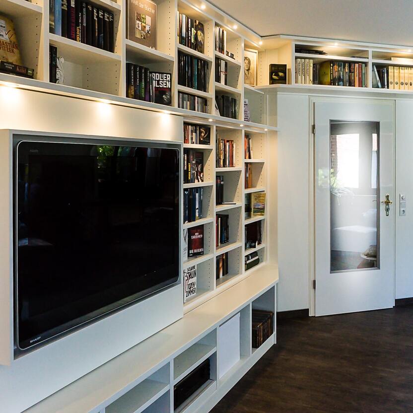 Maßgefertigte Medien- und Bücherwand mit integriertem TV Eine moderne, weiße Regalwand mit eingebautem Fernseher und zahlreichen Büchern, individuell angefertigt für eine stilvolle Wohnraumgestaltung.