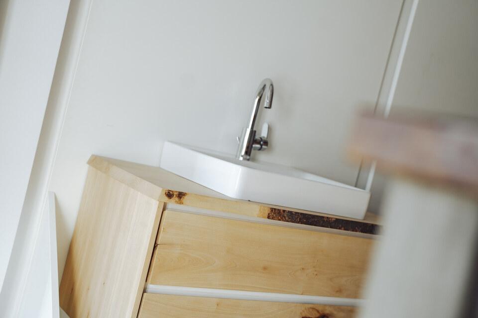 Badschrank aus hellem Holz mit einem weißen Waschbecken vor einer weißen Wand, in einem hellen Raum