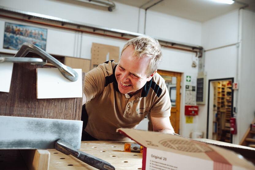 Ein lächelnder Tischler arbeitet mit einer Schraubzwinge an einem Holzstück in einer Werkstatt, um präzise Verbindungen herzustellen.