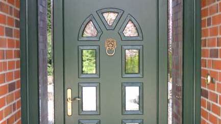 Dunkelgrüne Holztür mit Glaseinsätzen und Messinggriff Klassische dunkelgrüne Haustür aus Holz mit dekorativen Glaseinsätzen, Löwenkopf-Türklopfer und Messinggriff, eingerahmt von roten Klinkersteinen.