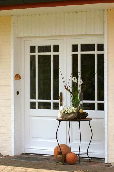 Stilvolle weiße Haustür aus Holz mit großen Glasfenstern, dekoriert mit Frühlingsblumen und Terrakottakugeln vor einem gelben Haus.