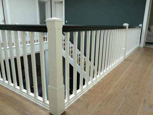 Großzügiger, heller Innenraum mit einem weißen Treppengeländer und einem schwarzen Handlauf auf einem Holzboden, vor einer dunkelgrauen Wand.