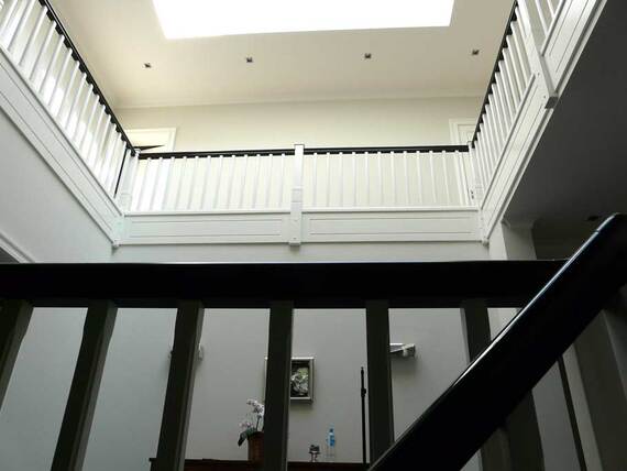 Blick nach oben auf ein helles Treppenhaus mit weißem Geländer und schwarzen Handläufen, umgeben von hohen Fenstern und einer luftigen Decke.