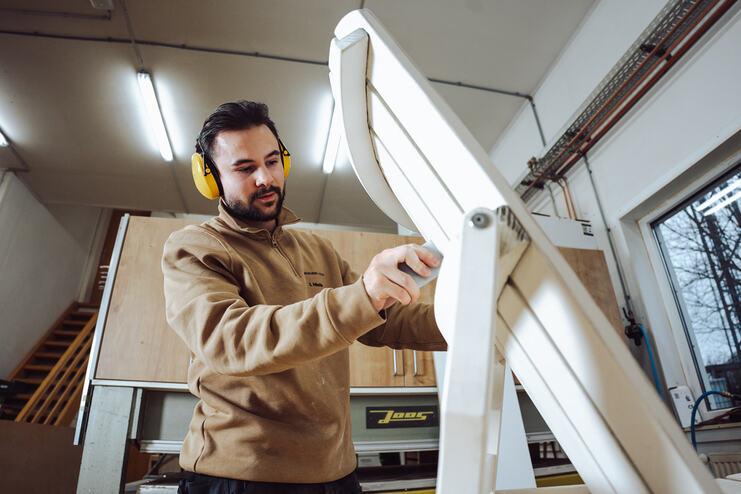Ein Tischler mit Geh&ouml;rschutz schleift einen wei&szlig;en Holzstuhl in einer Werkstatt, um eine saubere und pr&auml;zise Oberfl&auml;che zu erzielen.