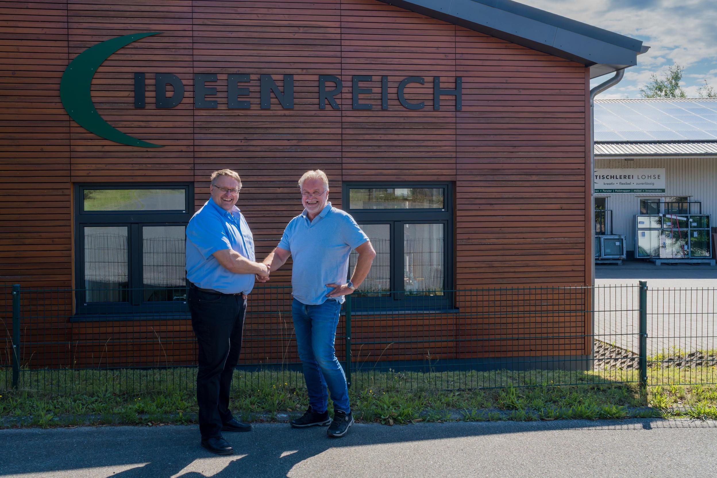 Lars Lohse und Thomas Jenning in blauen Hemden stehen vor dem IDEEN REICH, welches eine braune Holzfassade hat. Im Hintergrund ist ein Teil der Tischlerei Lohse abgebildet