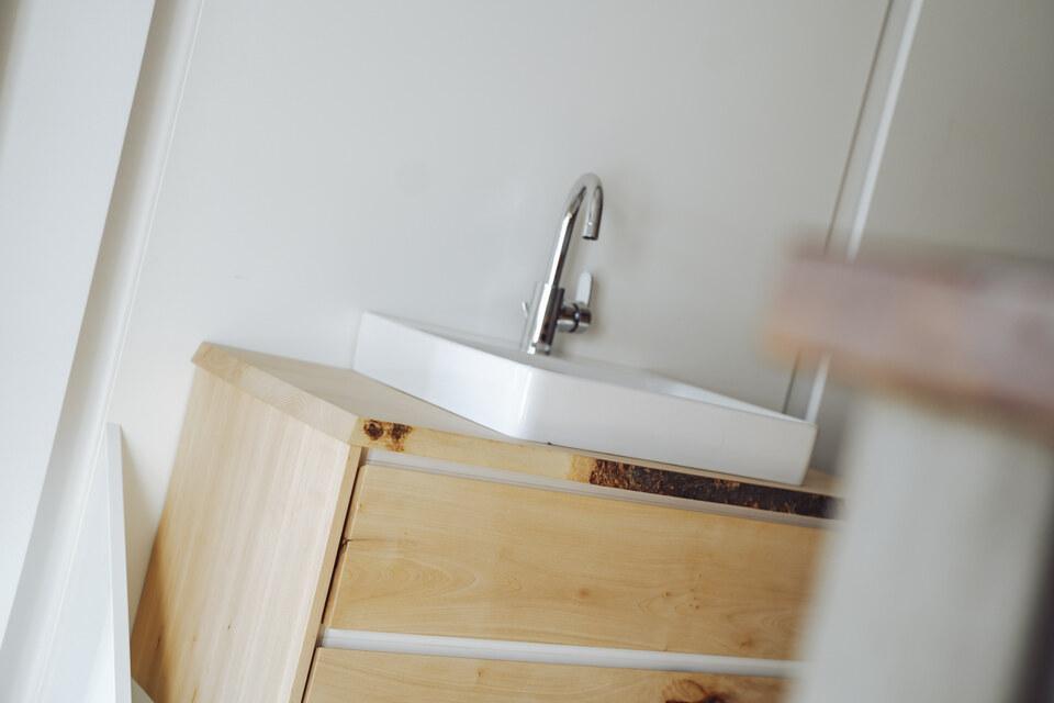 Badschrank im IDEEN REICH Badschrank aus hellem Holz mit einem weißen Waschbecken vor einer weißen Wand, in einem hellen Raum