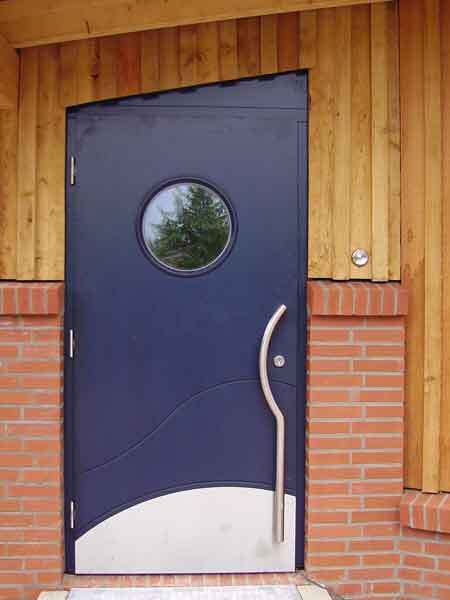 Dunkelblaue Eingangst&uuml;r mit rundem Fenster und geschwungenem Griff vor einer Holz- und Ziegelwand.