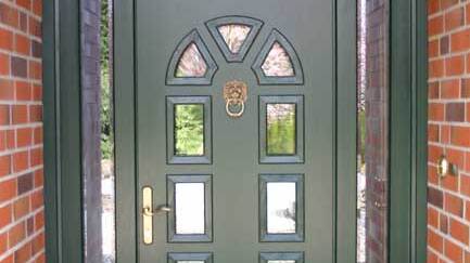 Dunkelgrüne Holztür mit Glaseinsätzen und Messinggriff Klassische dunkelgrüne Haustür aus Holz mit dekorativen Glaseinsätzen, Löwenkopf-Türklopfer und Messinggriff, eingerahmt von roten Klinkersteinen.